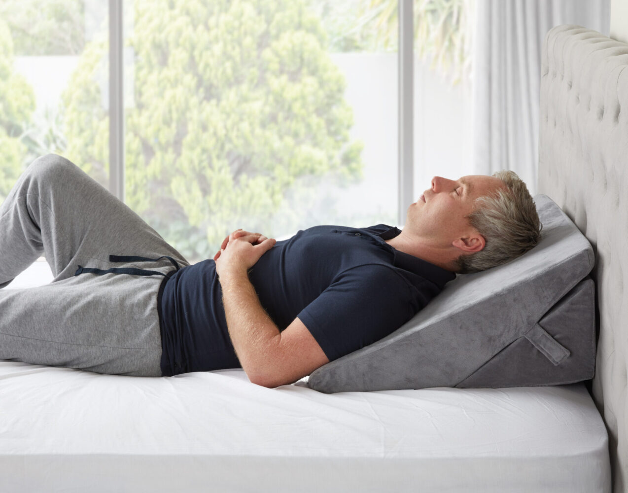 Man sleeping peacefully on the Slumbar wedge Pillow with white bedding and grey tracksuit bottoms