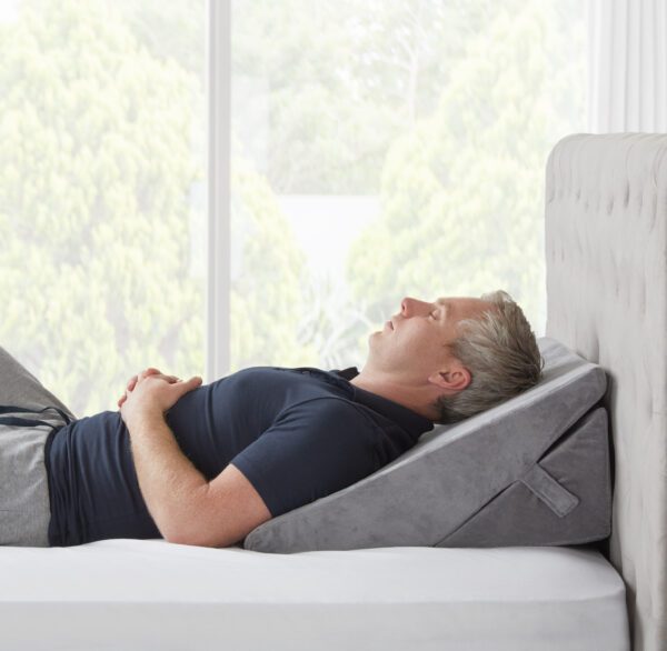 Man fast asleep on the Slumbar wedge pillow in its second highest level of incline, white bedding with garden in the background
