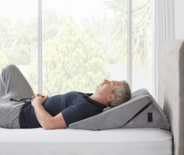 Man sleeping on the Slumbar Wedge pillow with it set at its lowest incline, White bed and lush green garden displayed in the background