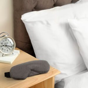 Slumbar sleep mask on table in bedroom, next to a grey bed with white cushions and an old alarm clock